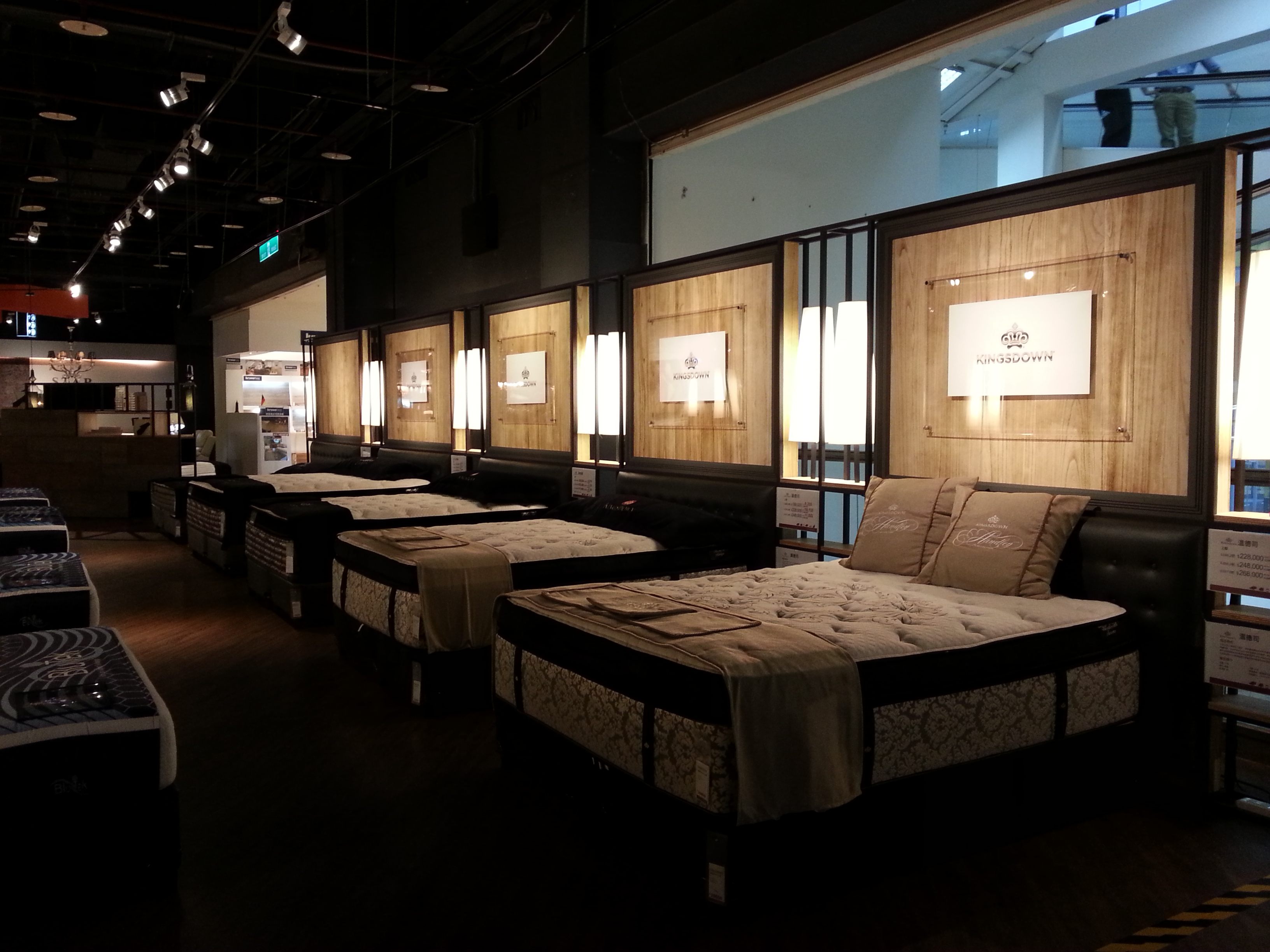 家具展厅内整齐陈列着多张KINGSDOWN品牌床垫,配备黑色皮质拉扣床头与米色绗缝床品。背景采用暖色木纹饰面墙及品牌Logo灯箱,右侧立牌标注“温德姆”系列及价