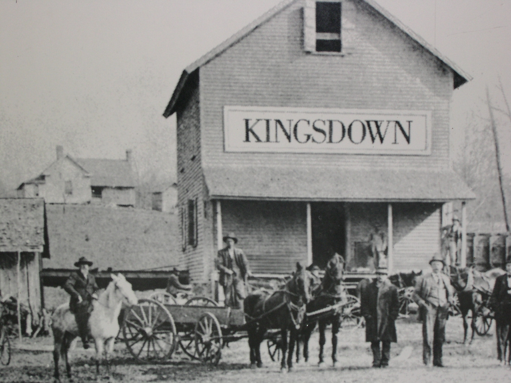 这是一张展现KINGSDOWN品牌早期历史的黑白照片。画面主体是一座宏伟的砖砌工厂建筑，正面醒目地悬挂着“KINGSDOWN”招牌。建筑前停放着多辆马车和运货平