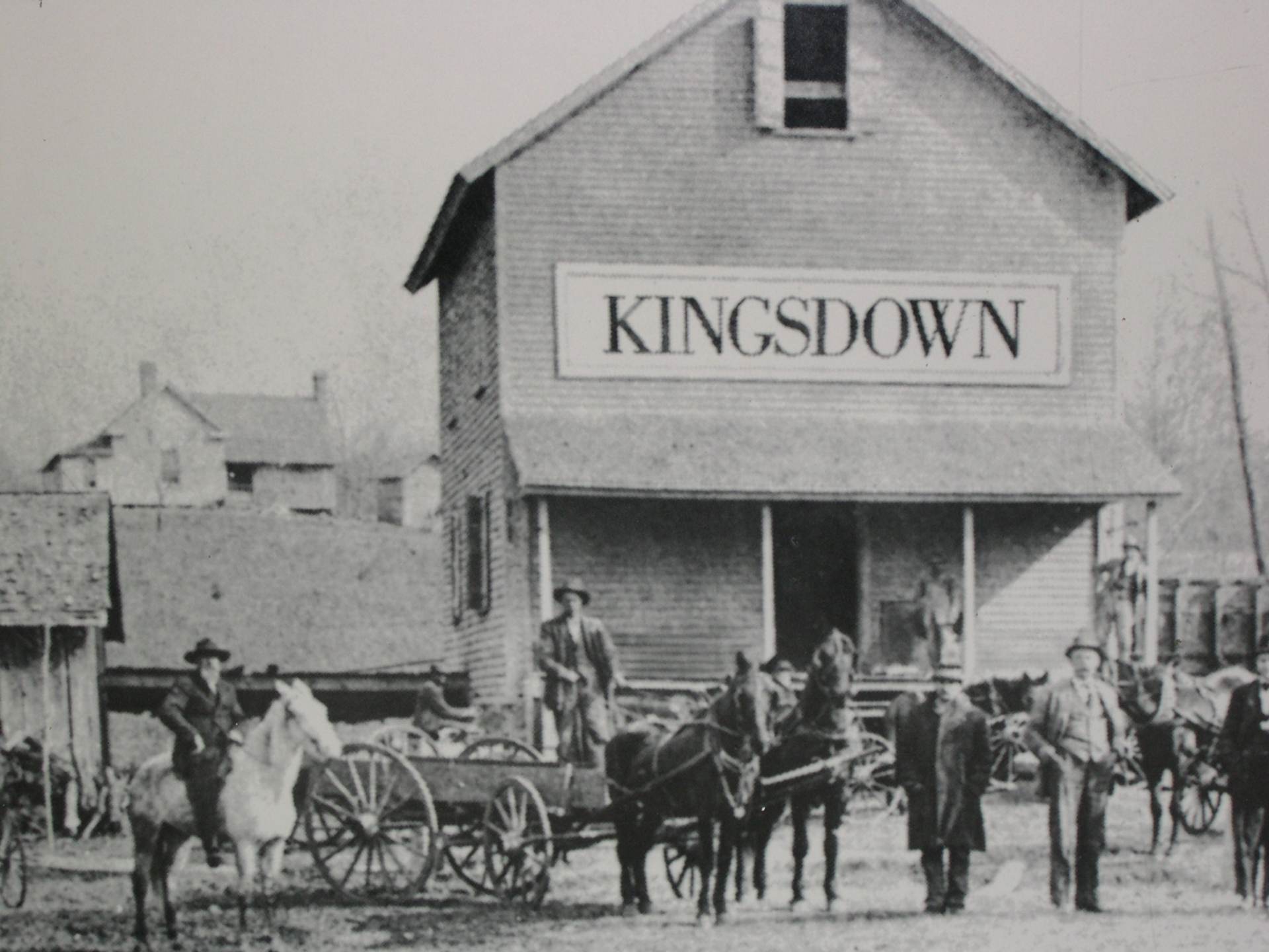 这是一张展现KINGSDOWN品牌早期历史的黑白照片。画面主体是一座宏伟的砖砌工厂建筑，正面醒目地悬挂着“KINGSDOWN”招牌。建筑前停放着多辆马车和运货平