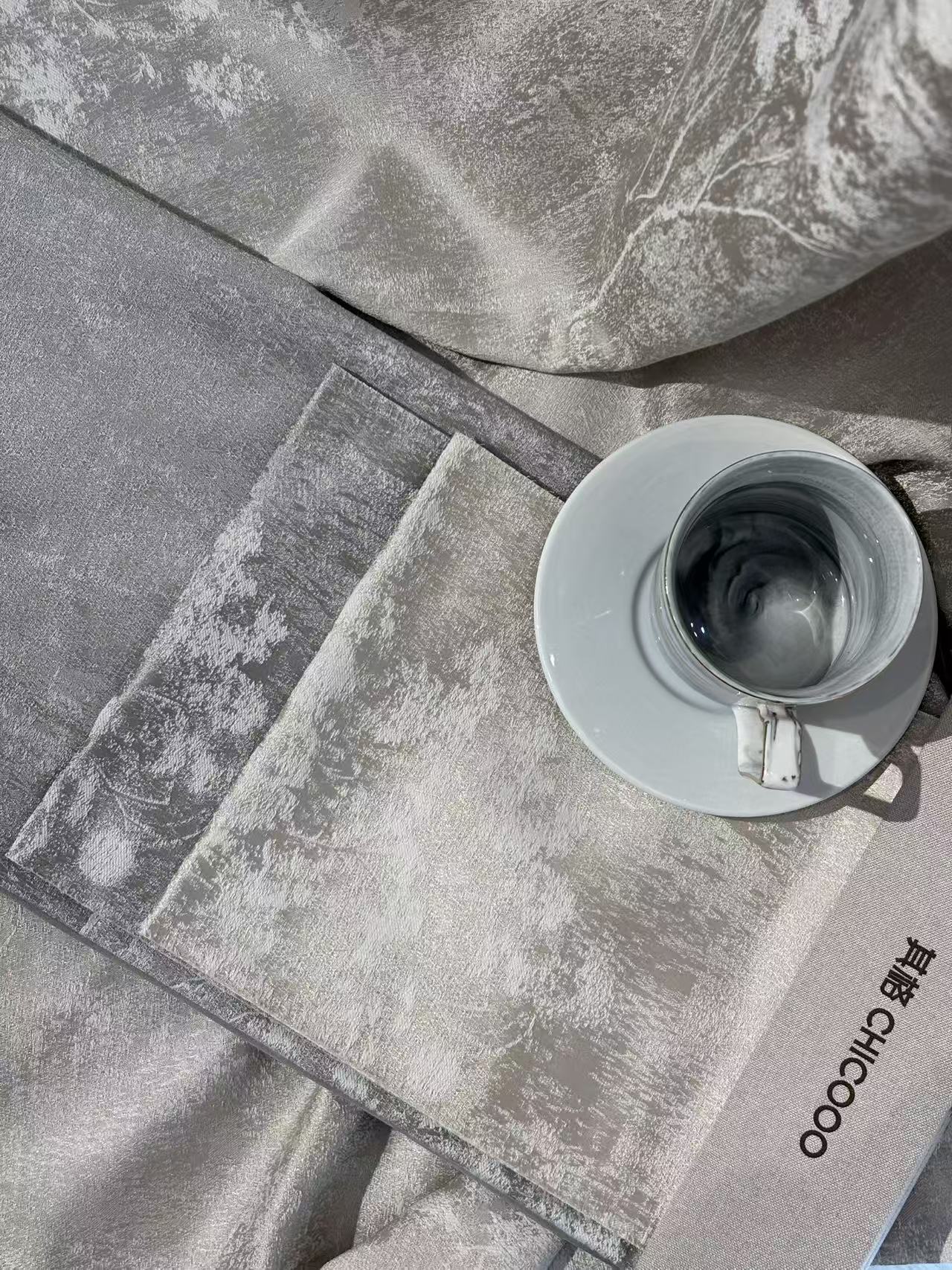 近距离特写展示了一款灰色调的家居纺织品，表面呈现出细腻的大理石纹理或云雾状渐变，质感柔软且带有微光泽，疑似丝绒或高支棉材质。画面右侧搭配白色咖啡杯碟，营造出精致
