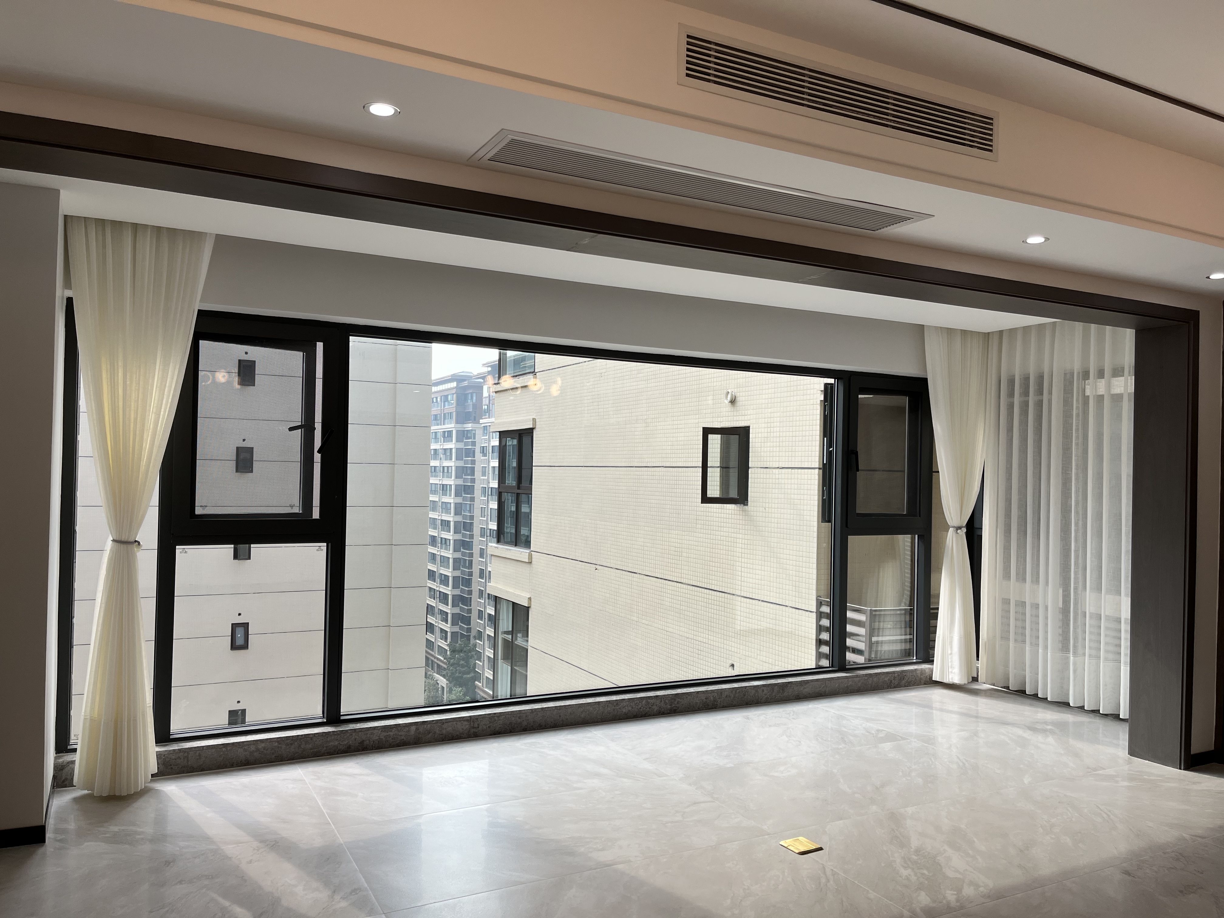 现代简约风格客厅封阳台实景,核心展示黑色断桥铝落地窗,采用大固定玻璃搭配两侧开启扇,视野开阔采光充足。窗边悬挂白色纱帘,地面铺设灰色亮面瓷砖,顶部集成中央空调与