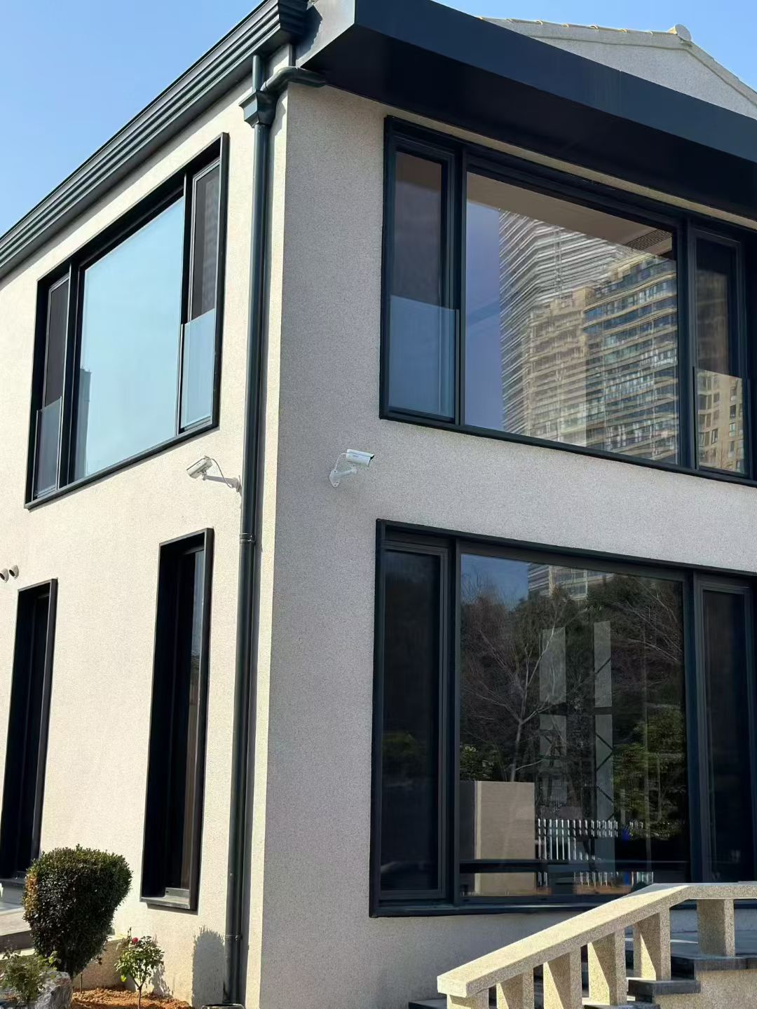 现代风格别墅或自建房外立面实拍,墙面采用米白色真石漆,质感细腻均匀。搭配黑色断桥铝大落地窗及黑色檐口落水管,形成经典的黑白极简配色。大面积玻璃窗不仅采光极佳,还