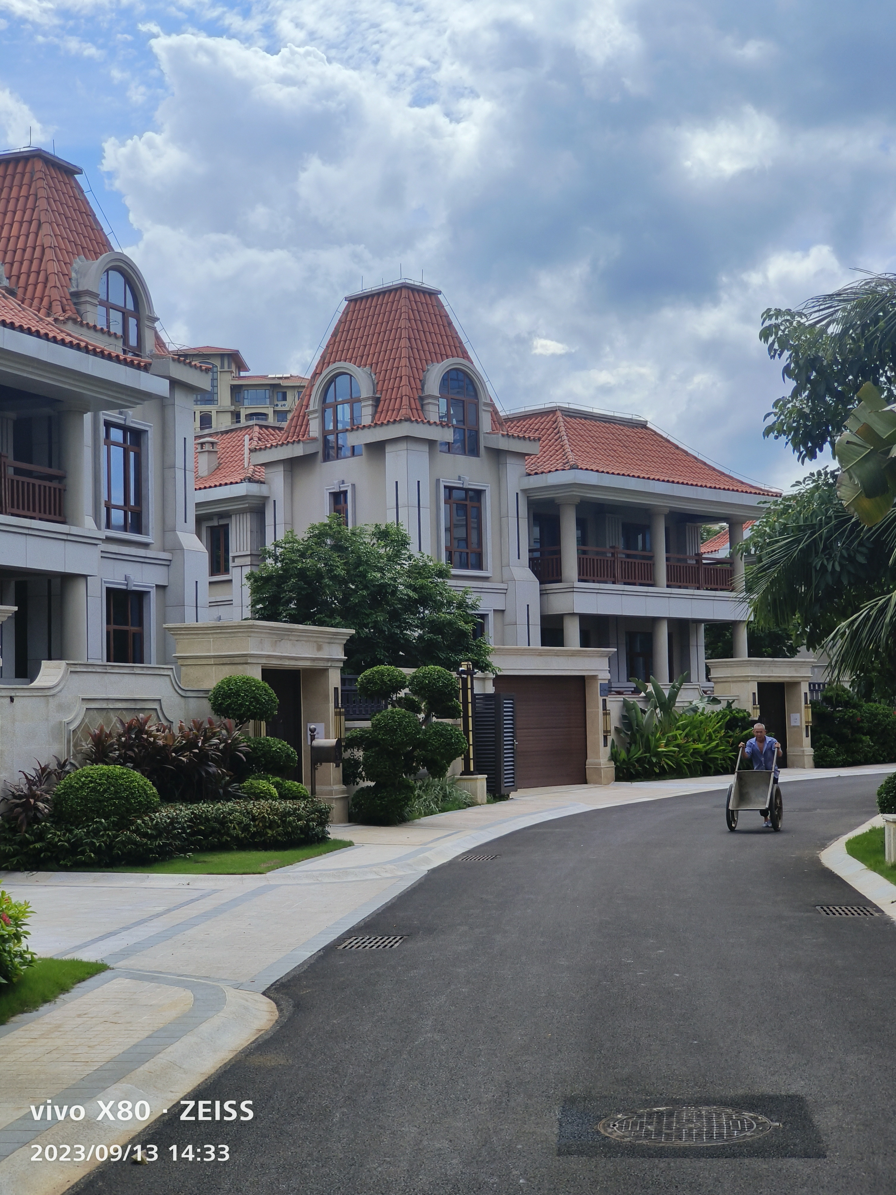这是一张展示高档欧式别墅区的实景照片。画面中矗立着多栋米白色外墙的独栋别墅,搭配醒目的红色坡屋顶和拱形落地窗,尽显欧式古典风情。建筑采用石材干挂工艺,质感细腻,