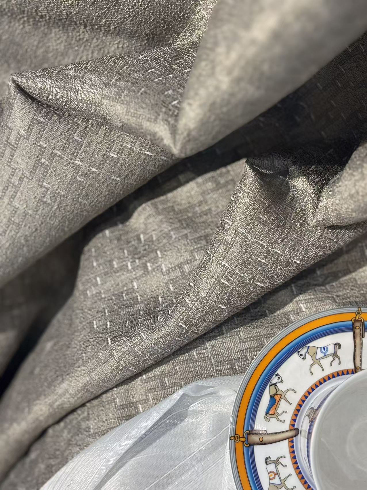 这是一张家居软装面料的特写图，主体为银灰色提花窗帘布料，表面带有细腻的十字纹理和微光泽感，质感厚实且垂坠自然。搭配下方透光的白色窗纱，营造出层次丰富的视觉效果。