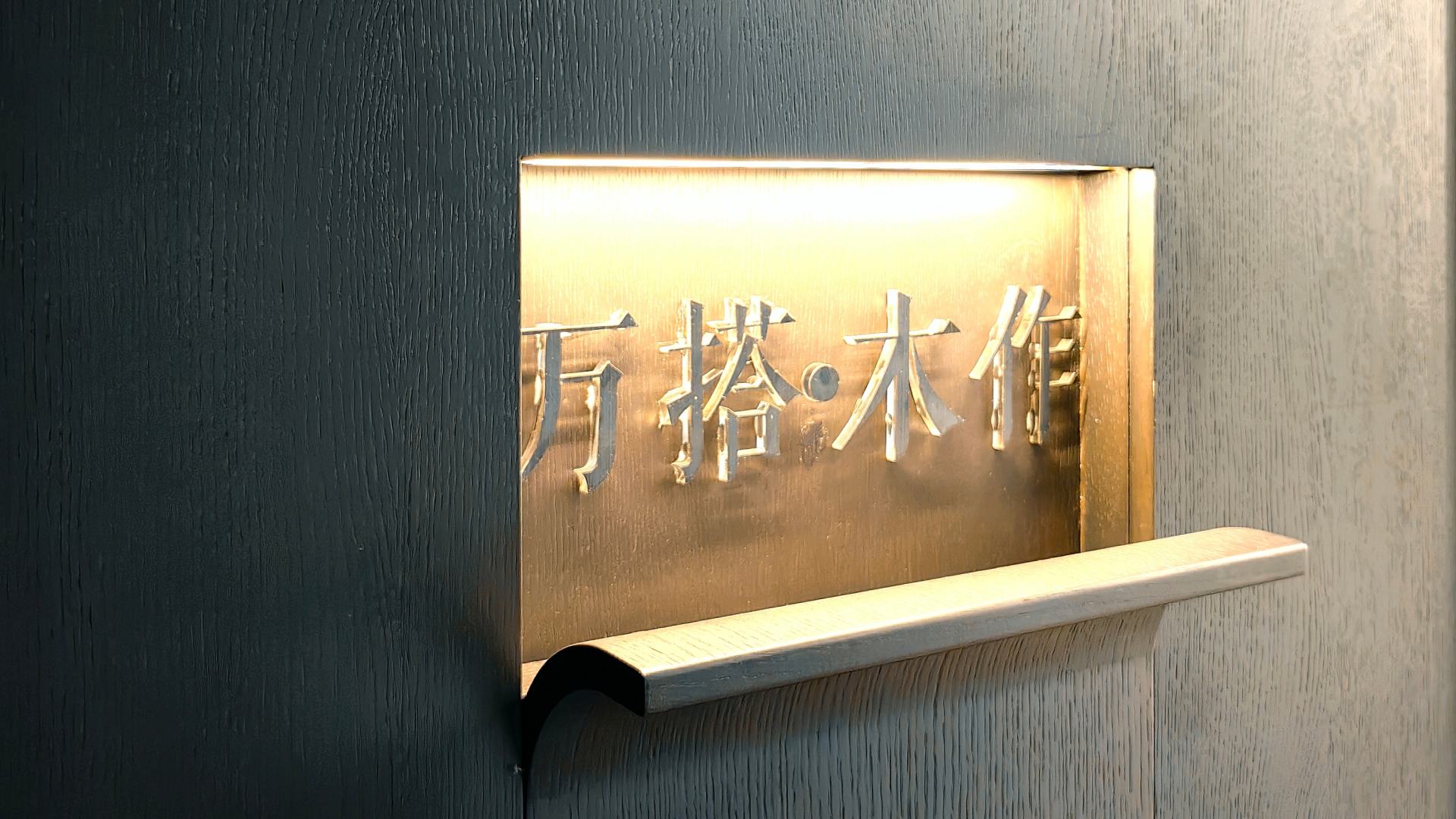 深色垂直木纹背景墙面上，嵌入安装有一块金色拉丝金属标识牌，顶部暖光投射凸显立体字“万搭·木作”。牌体下方延伸出浅色原木弧形搁板，兼具品牌展示与轻量收纳功能。整体