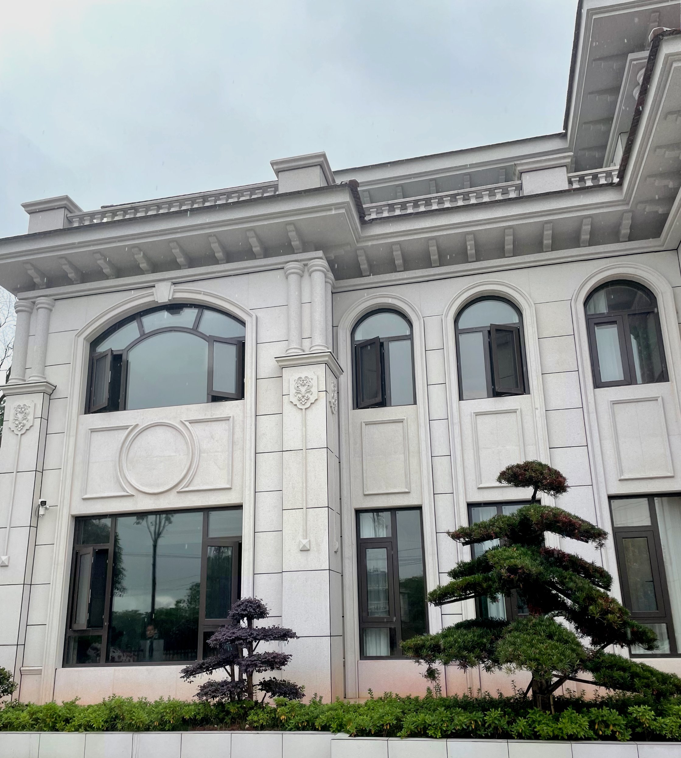 这是一张简欧风格别墅外立面的实景拍摄。建筑外墙采用白色石材或高品质仿石漆，搭配深灰色断桥铝拱形大窗，视觉对比鲜明。立面设计包含罗马柱、精致浮雕与圆形装饰线条，工