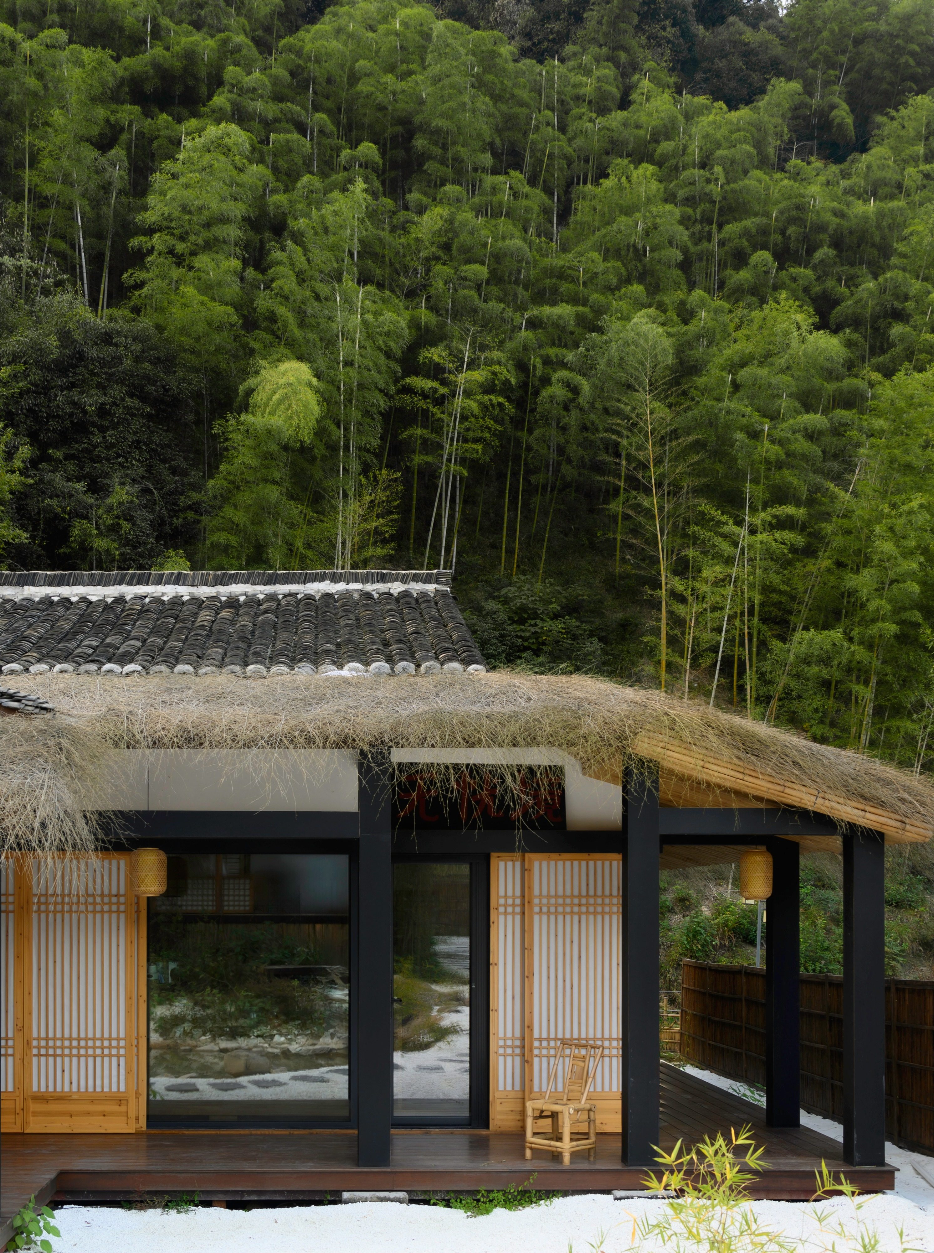 位于茂密竹林中的传统风格建筑，融合茅草屋顶与黑色瓦片设计，木质格栅门窗搭配黑色框架结构，营造自然质朴氛围。建筑前设有木质平台和白色碎石地面，配以竹编灯笼和竹制座